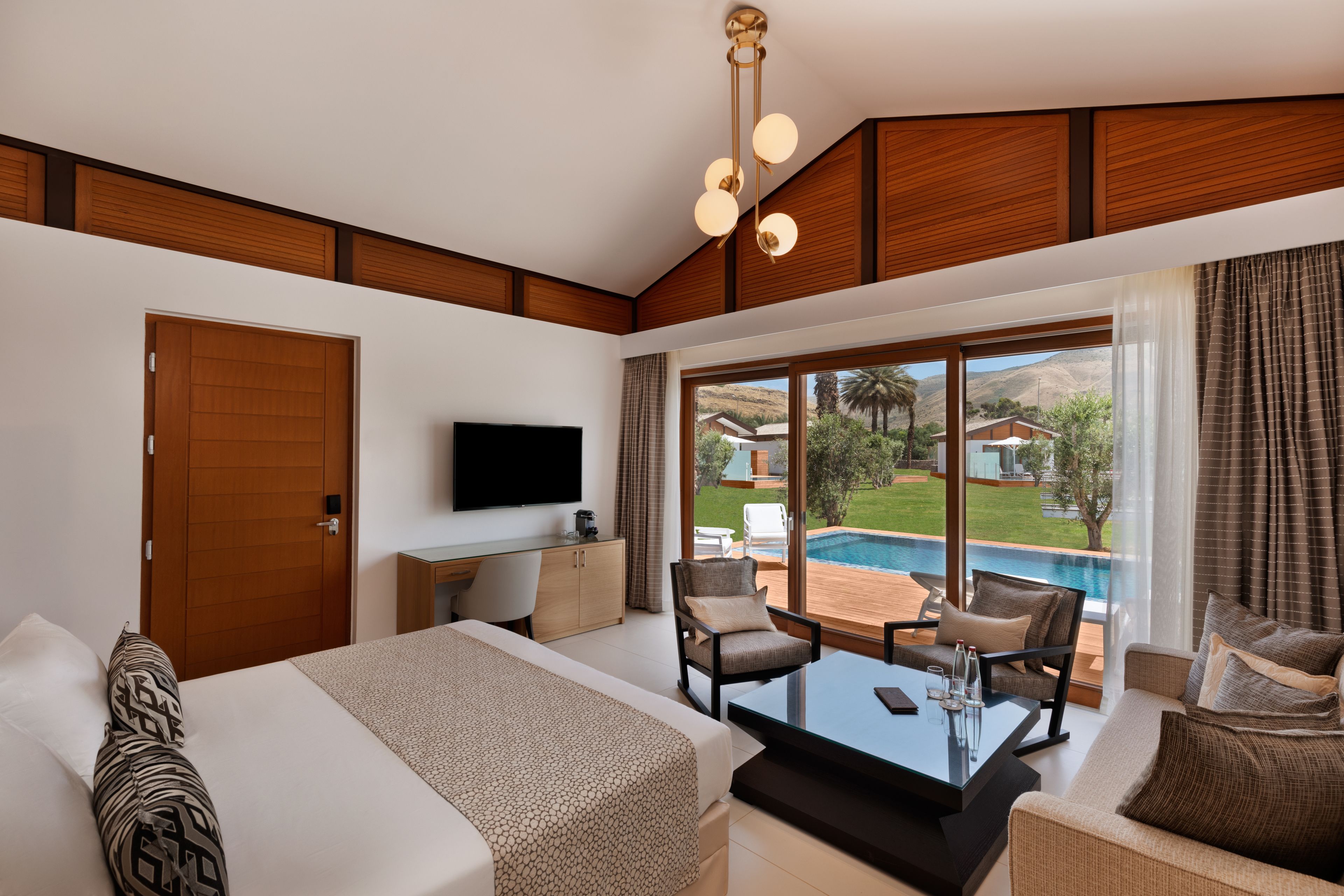 A modern hotel room with a large bed, a desk, and a seating area with a view of a swimming pool and mountains.