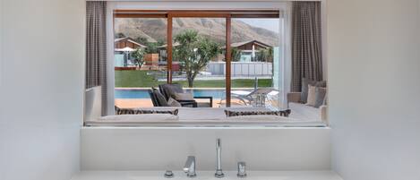 A modern bathroom with a large bathtub, a window overlooking a pool and outdoor seating area.