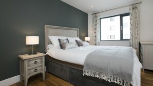 Un dormitorio con cama, mesitas de noche, ventana con cortinas y vista a edificios.