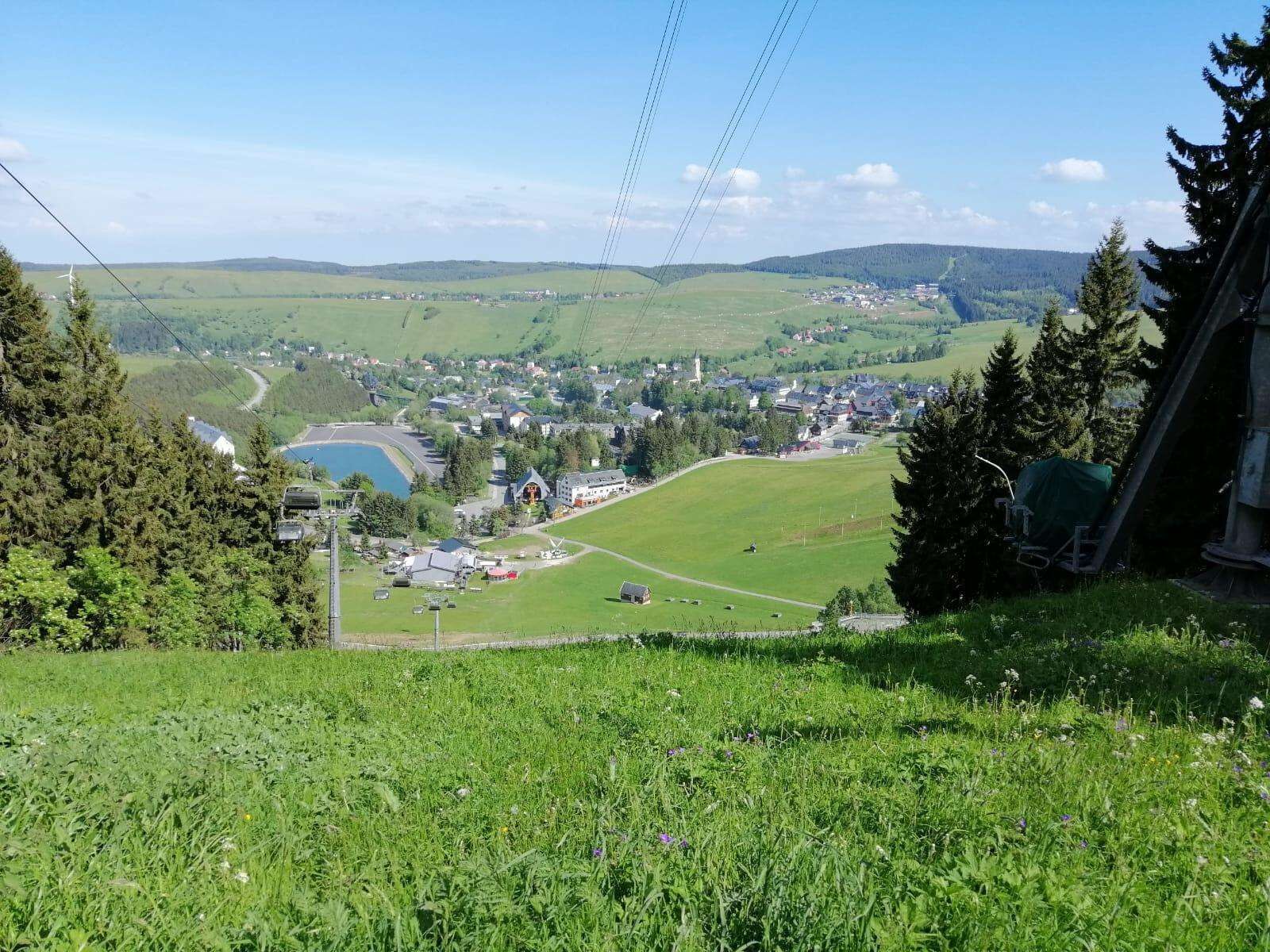 A panoramic view of a town with a lake, surrounded by green hills and trees.