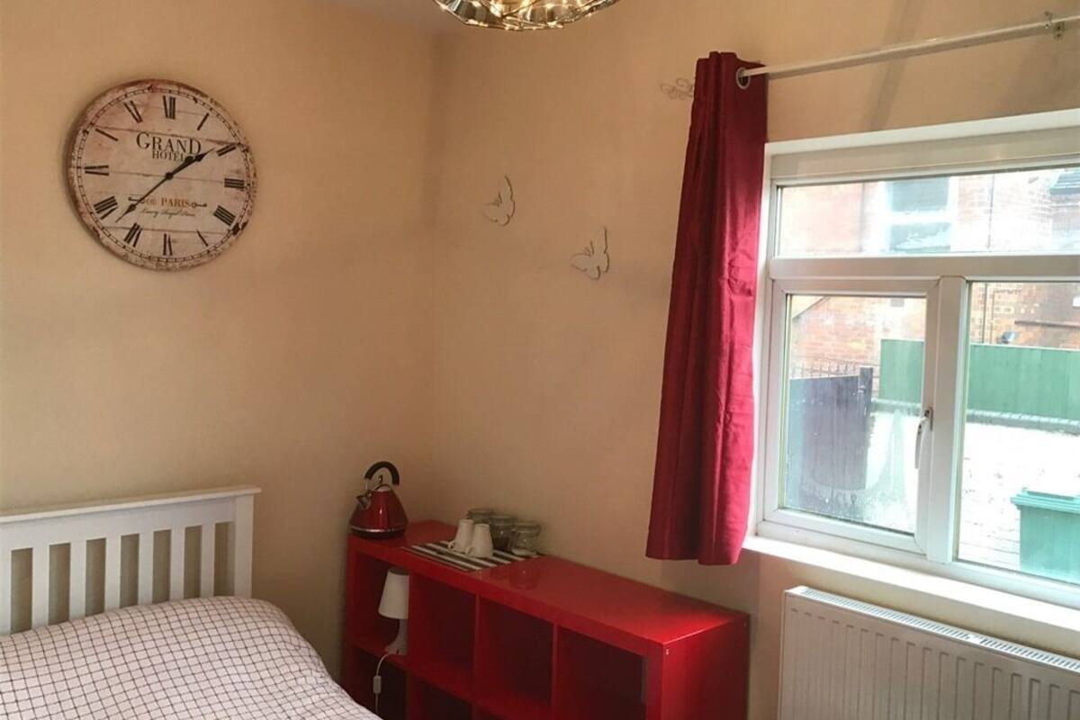 A bedroom with a bed, a red nightstand, a wall clock, and a window with a view of the outside.