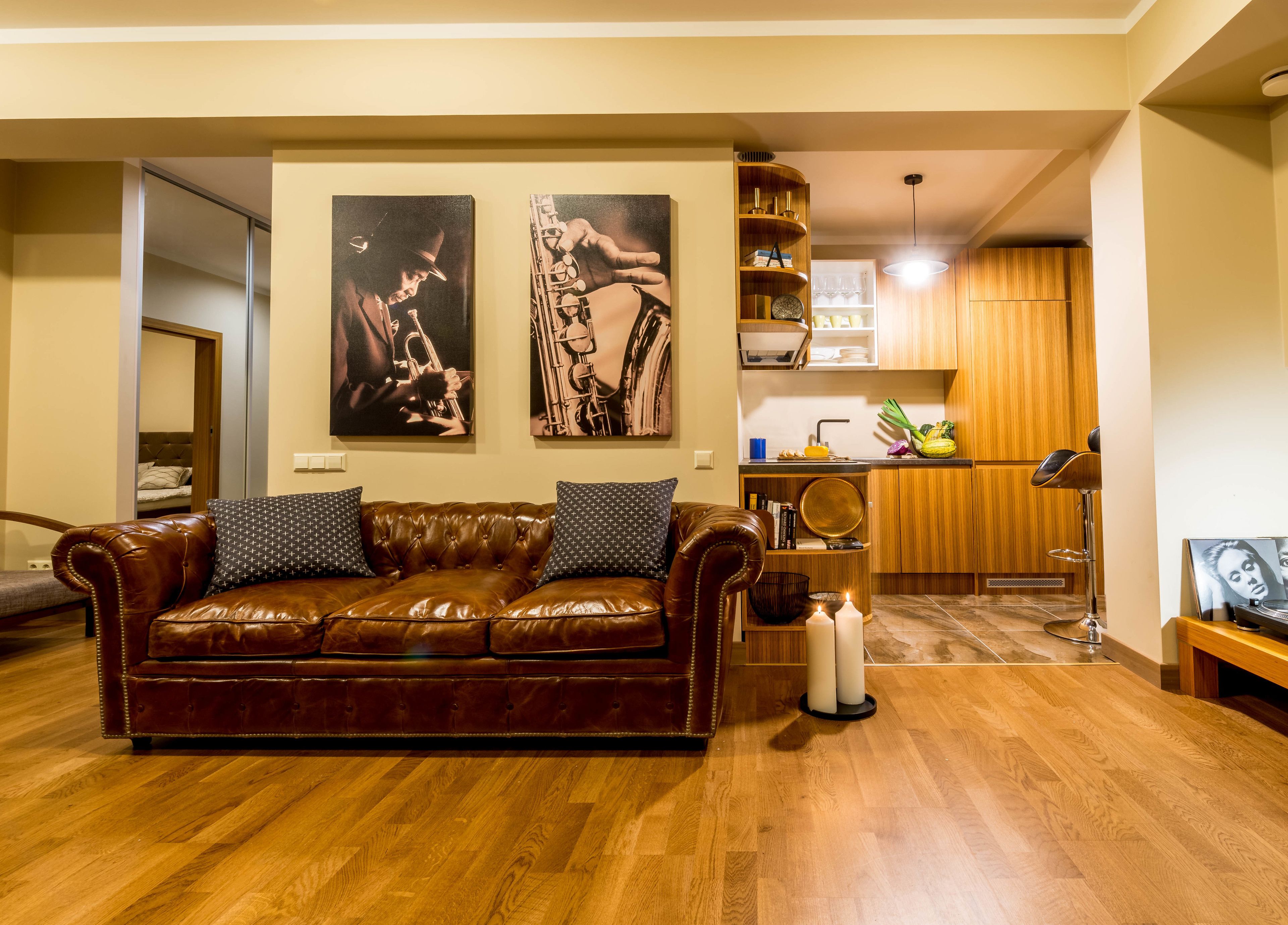 A living room with a brown leather sofa, two wall-mounted paintings, a wooden floor, and a kitchen area with wooden cabinets and a bar stool.