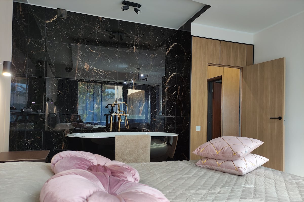 Ein modernes Schlafzimmer mit Marmorwand, einem runden Spiegel, einer Holztür und einem Bett mit rosa Kissen.