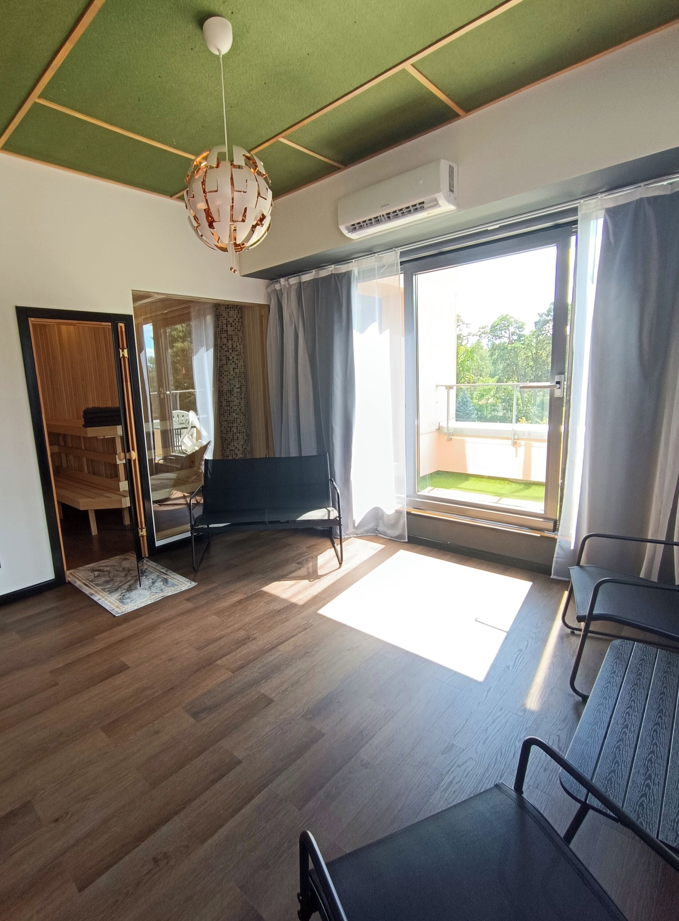 Ein geräumiges Zimmer mit Holzboden, Deckenventilator, großem Fenster mit Vorhängen und Blick ins Freie.
