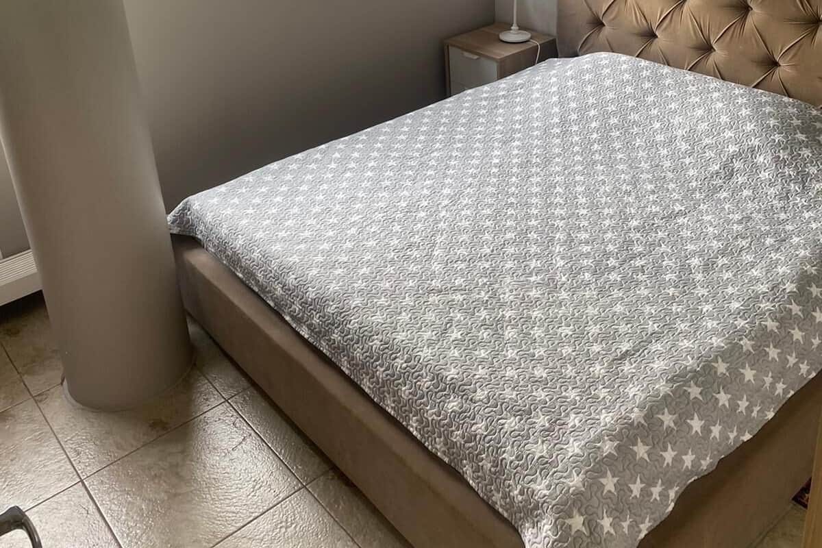 Ein Schlafzimmer mit Bett, Nachttisch, Lampe, Fenster mit Blick auf Grünflächen und einem Bild an der Wand.