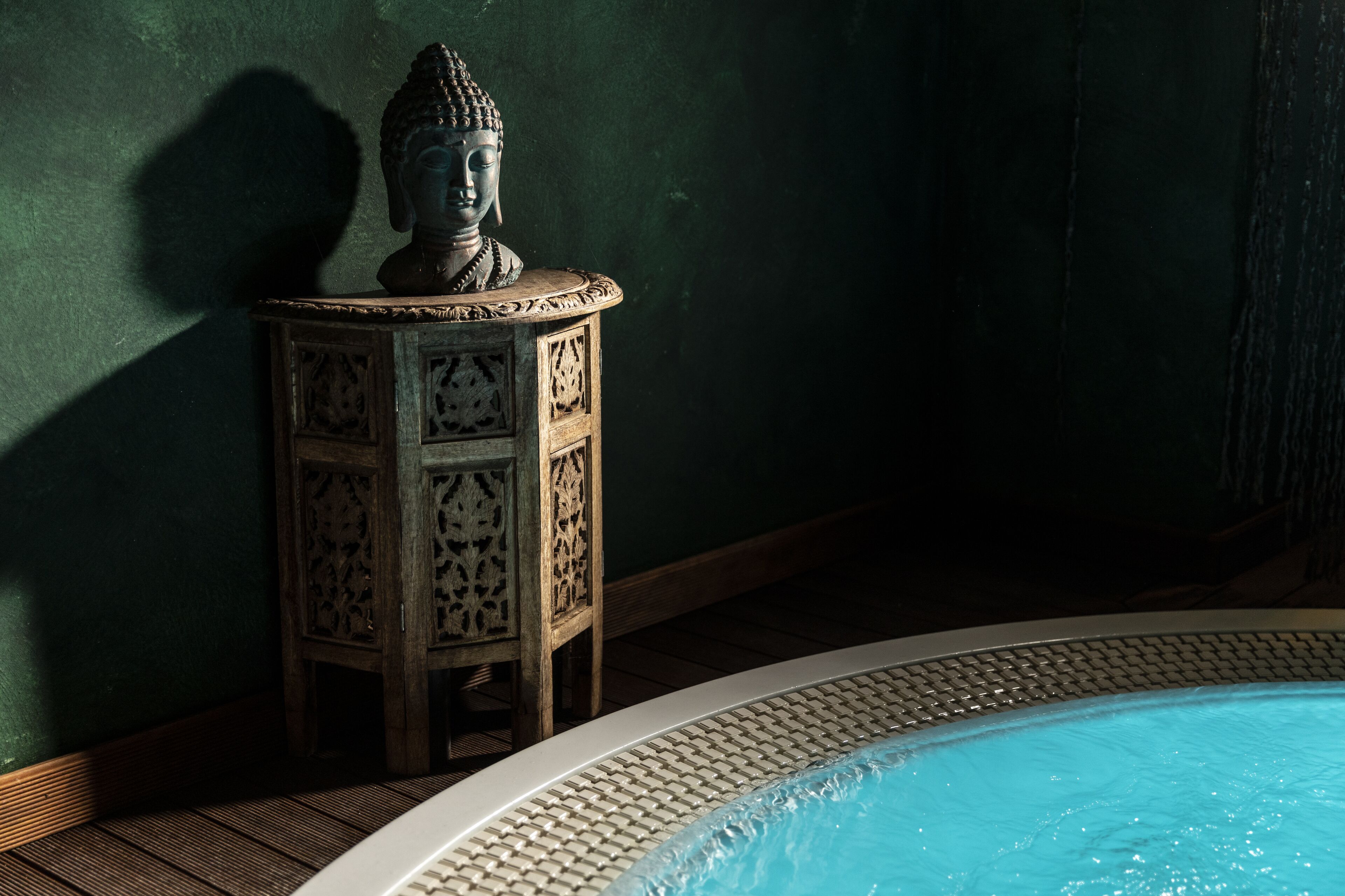 Una pequeña mesa de madera, finamente tallada, con una estatua de Buda encima, ubicada junto a una piscina con borde de azulejos.