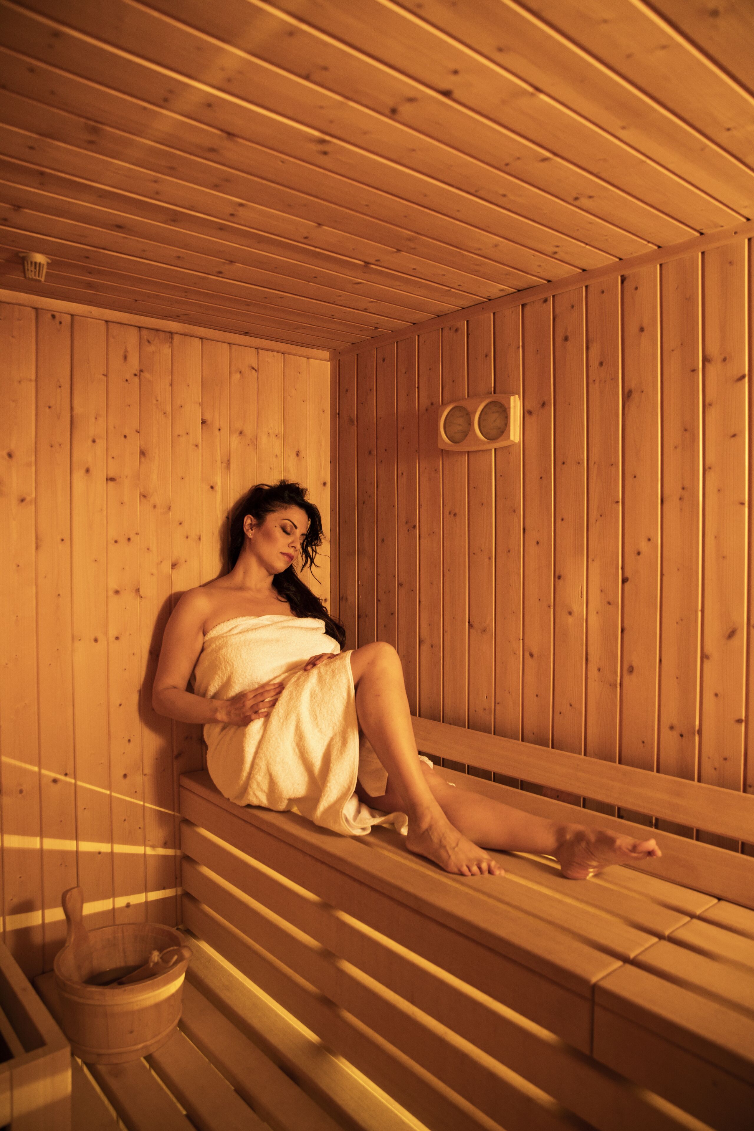 Una sauna con paredes y bancos de madera, un cubo de madera y una persona envuelta en una toalla.