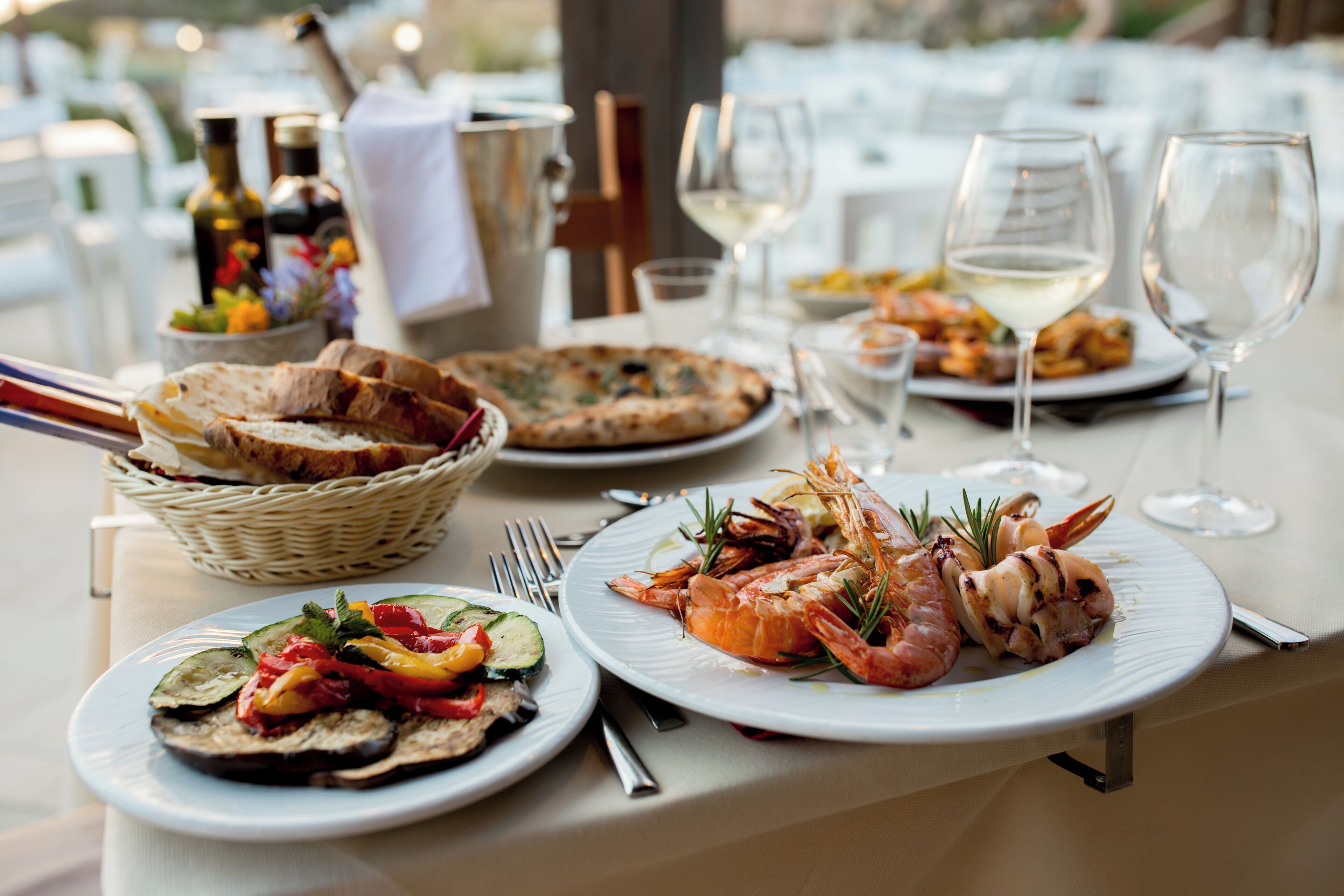 A dining table set with plates of seafood, bread, and a pizza, along with wine glasses and bottles.