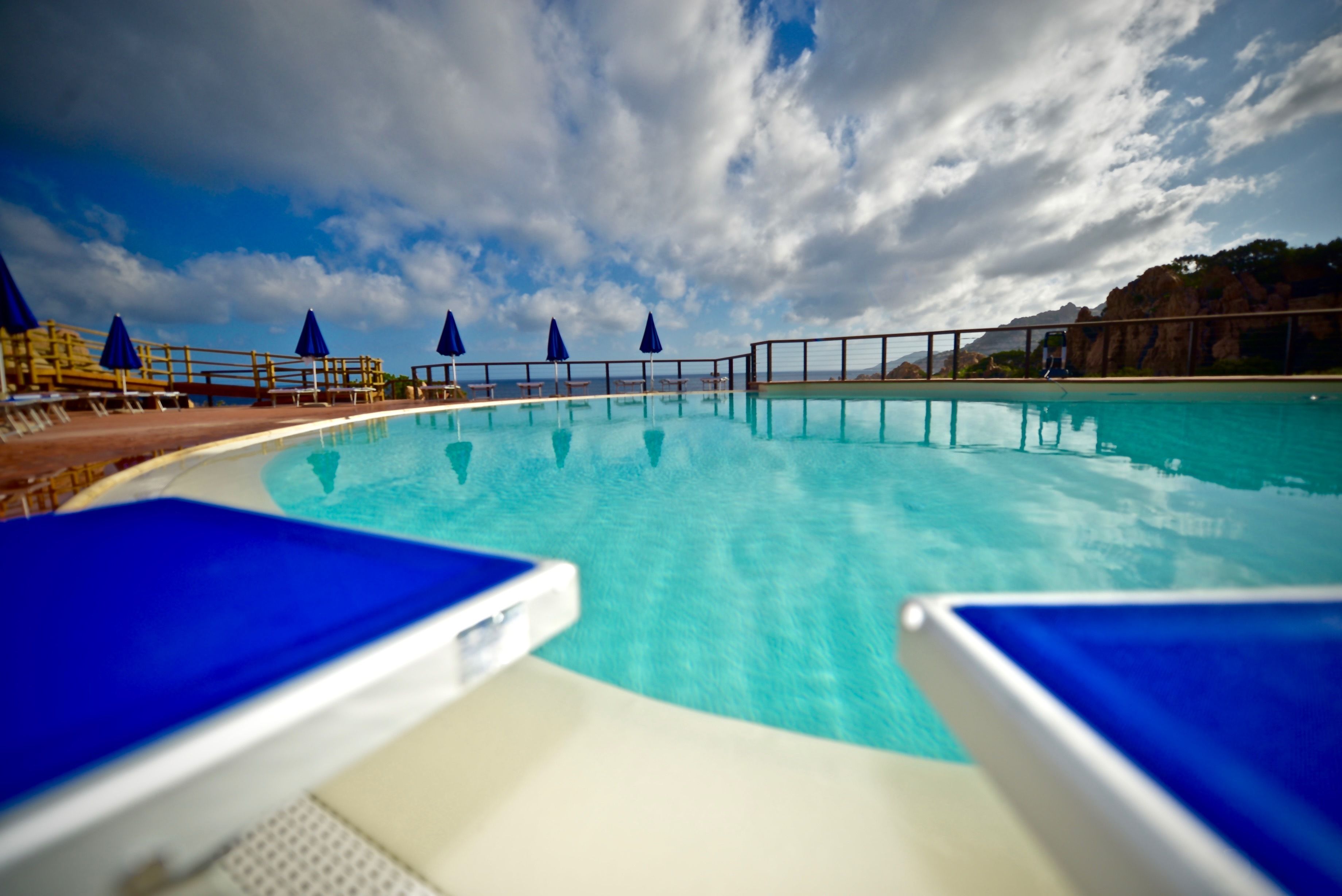 Una piscina con sillones reclinables azules y sombrillas, rodeada por una terraza de madera y una barandilla.