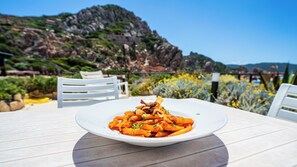 A white plate of pasta with a tomato-based sauce, served on a white table with a view of a rocky landscape and yellow flowers in the background. - Hotel Costa Paradiso (Trinità d'Agultu e Vignola)