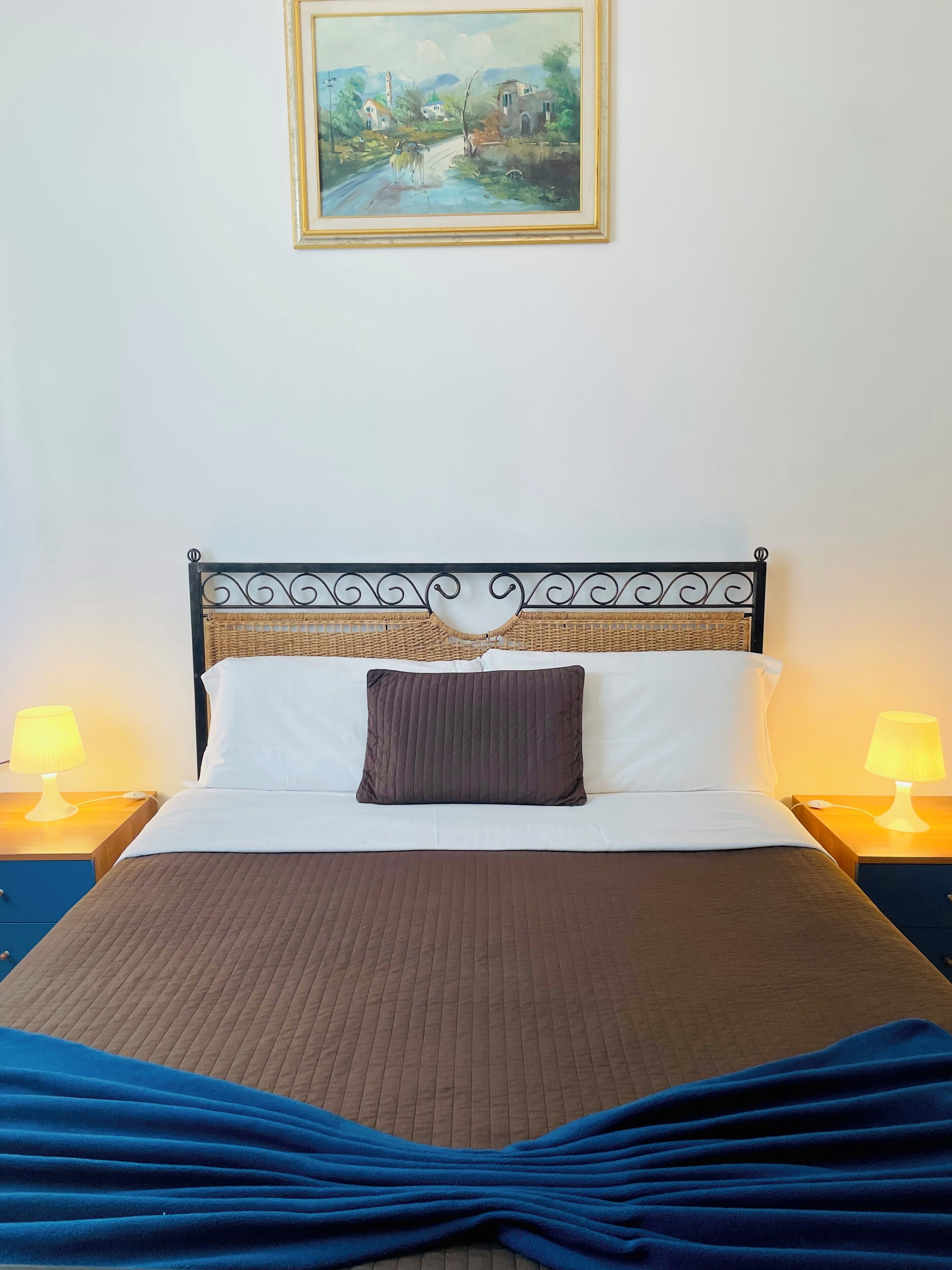 A neatly made bed with a brown bedspread, a single brown pillow, and a blue blanket. There are two bedside lamps with yellow shades, and a painting of a landscape hangs on the wall above the bed.