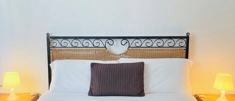 A neatly made bed with a brown bedspread, a single brown pillow, and a blue blanket. There are two bedside lamps with yellow shades, and a painting of a landscape hangs on the wall above the bed.
