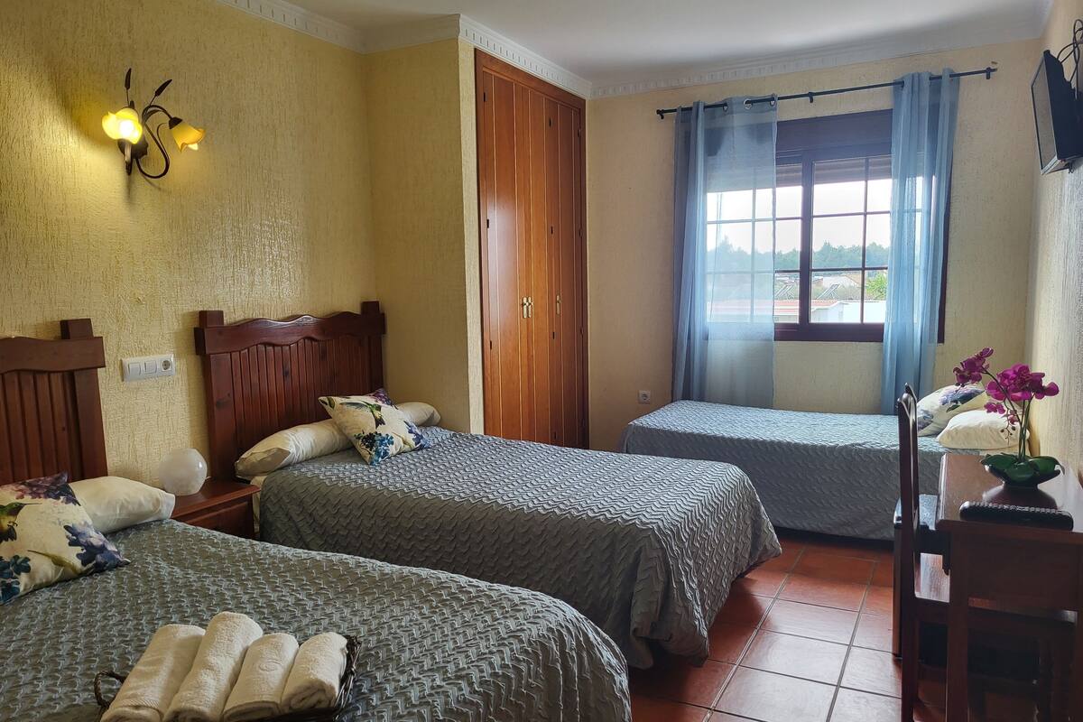A hotel room with two beds, a wooden wardrobe, a desk with a chair, a window with curtains, and a wall-mounted light fixture.