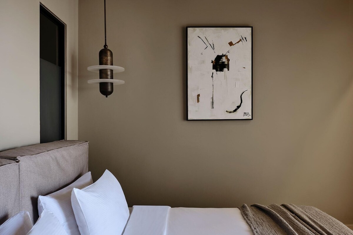 Een hotelkamer met een bed, kussens, een hanglamp en een abstract schilderij in een lijst aan de muur.