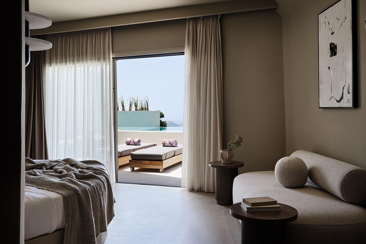 Een hotelkamer met een bed, een bank, een tafeltje en uitzicht op een zwembad en groen.