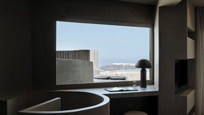 A modern hotel room with a large window, a curved desk, a chair, a flat-screen TV, and a view of the coastline.