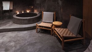 A dimly lit room with a large bathtub, two wooden chairs, and a small round table.