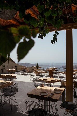 Un área de comedor al aire libre con mesas y sillas, vista al mar y una pérgola con plantas.