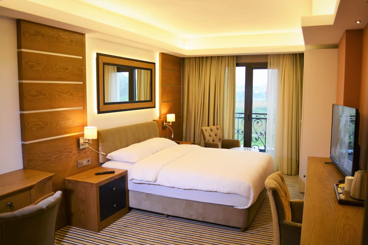 A hotel room with a large bed, a desk with a TV, a chair, and a balcony with curtains.