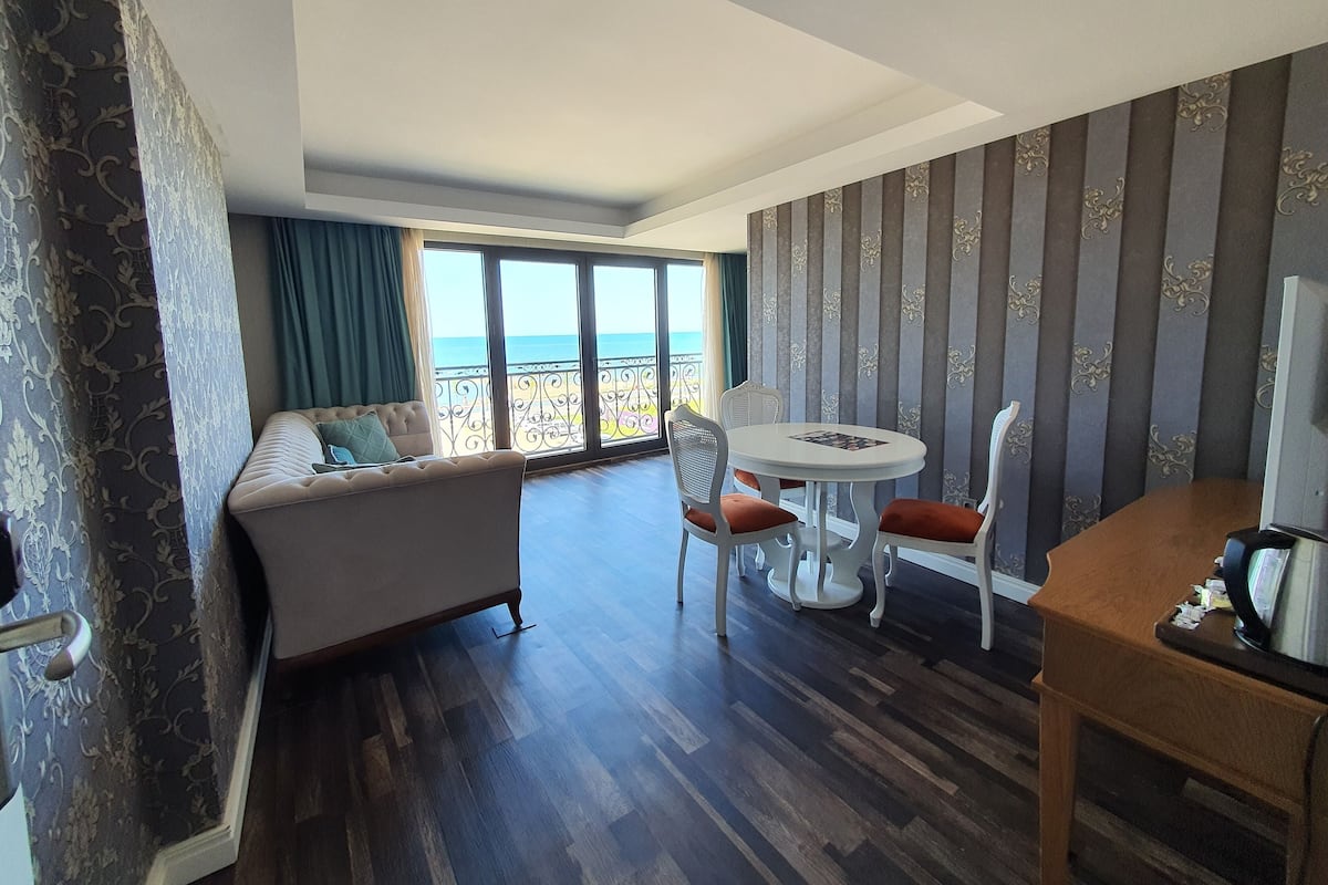 A hotel room with a balcony, a sofa, a dining table with chairs, and a TV.