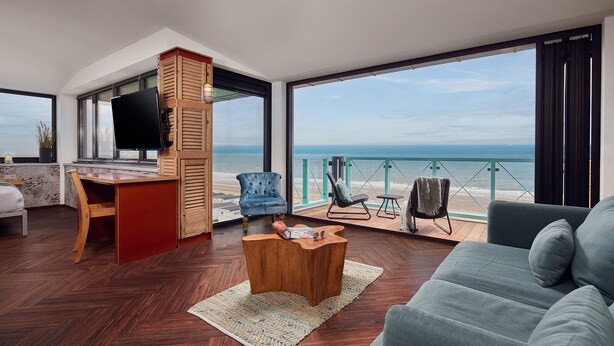 A modern living room with a large window overlooking the beach, a wooden desk, a blue armchair, and a grey sofa. - Beachhouse Hotel (Zandvoort)