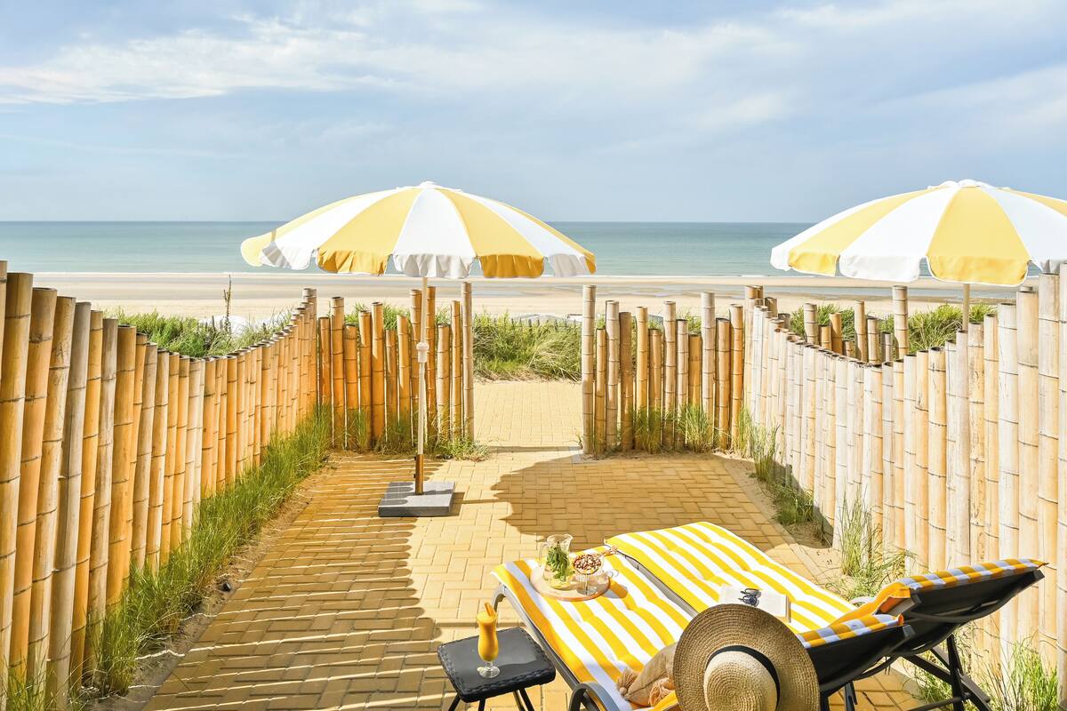 Een loungehoek aan het strand met twee ligbedden, een tafel met drankjes en twee grote parasols voor schaduw.