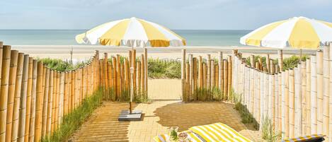 Eine Strandlounge mit zwei Liegen, einem Tisch mit Getränken und zwei großen Sonnenschirmen, die Schatten spenden.