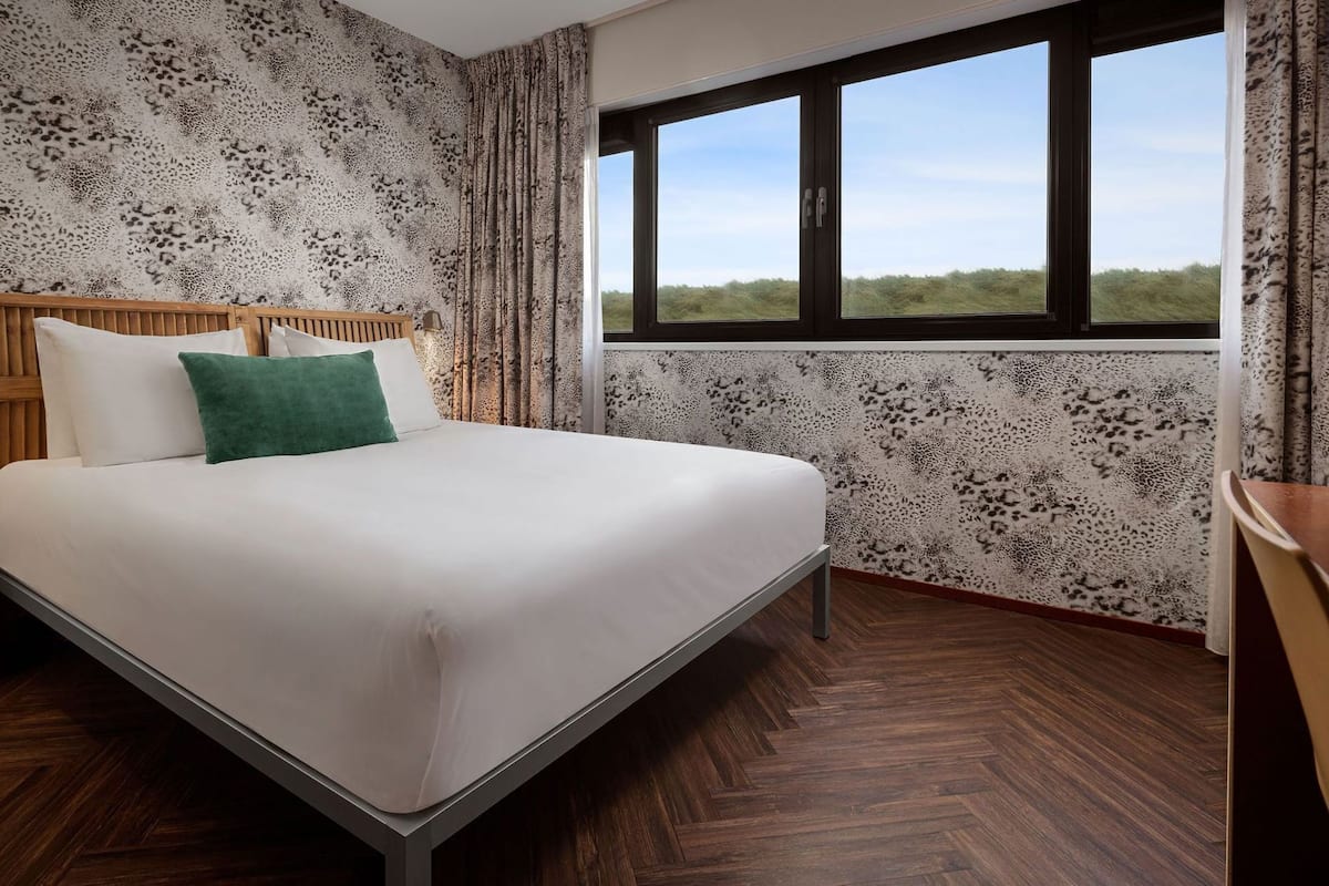 Een slaapkamer met een bed, een houten hoofdbord, een raam met uitzicht en behang met een patroon.