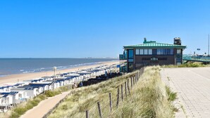 A coastal building with a green roof, adjacent to a beach with rows of white beach houses, a paved walkway, and a grassy embankment. - Beachhouse Hotel (Zandvoort)