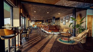 A modern bar area with wooden bar counter, bar stools, and wicker chairs. - Beachhouse Hotel (Zandvoort)