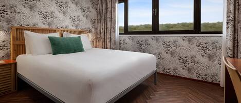 A neatly made bed with white linens and a green pillow, a wooden headboard, and a window with a view of greenery.