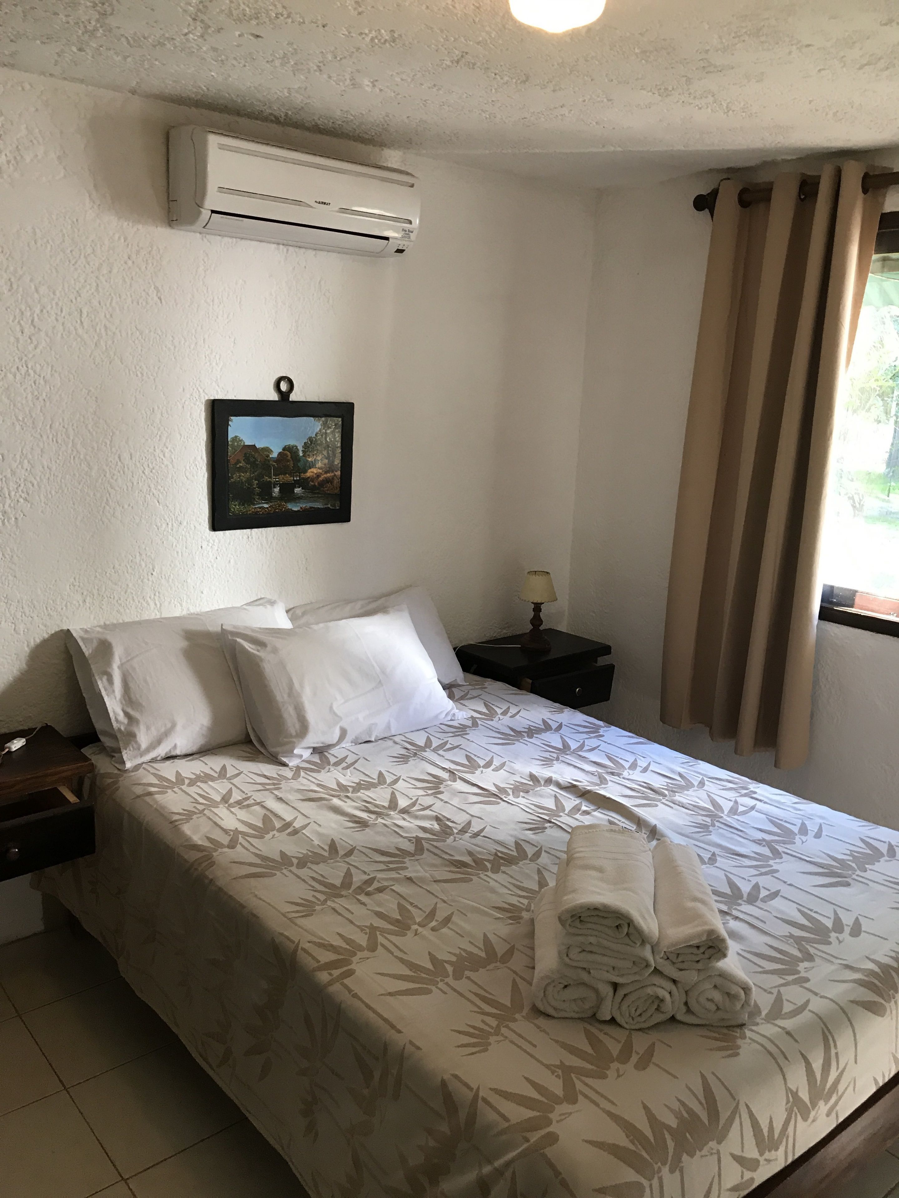 A neatly made bed with a floral bedspread, a wall-mounted air conditioner, a framed picture, and a window with curtains.