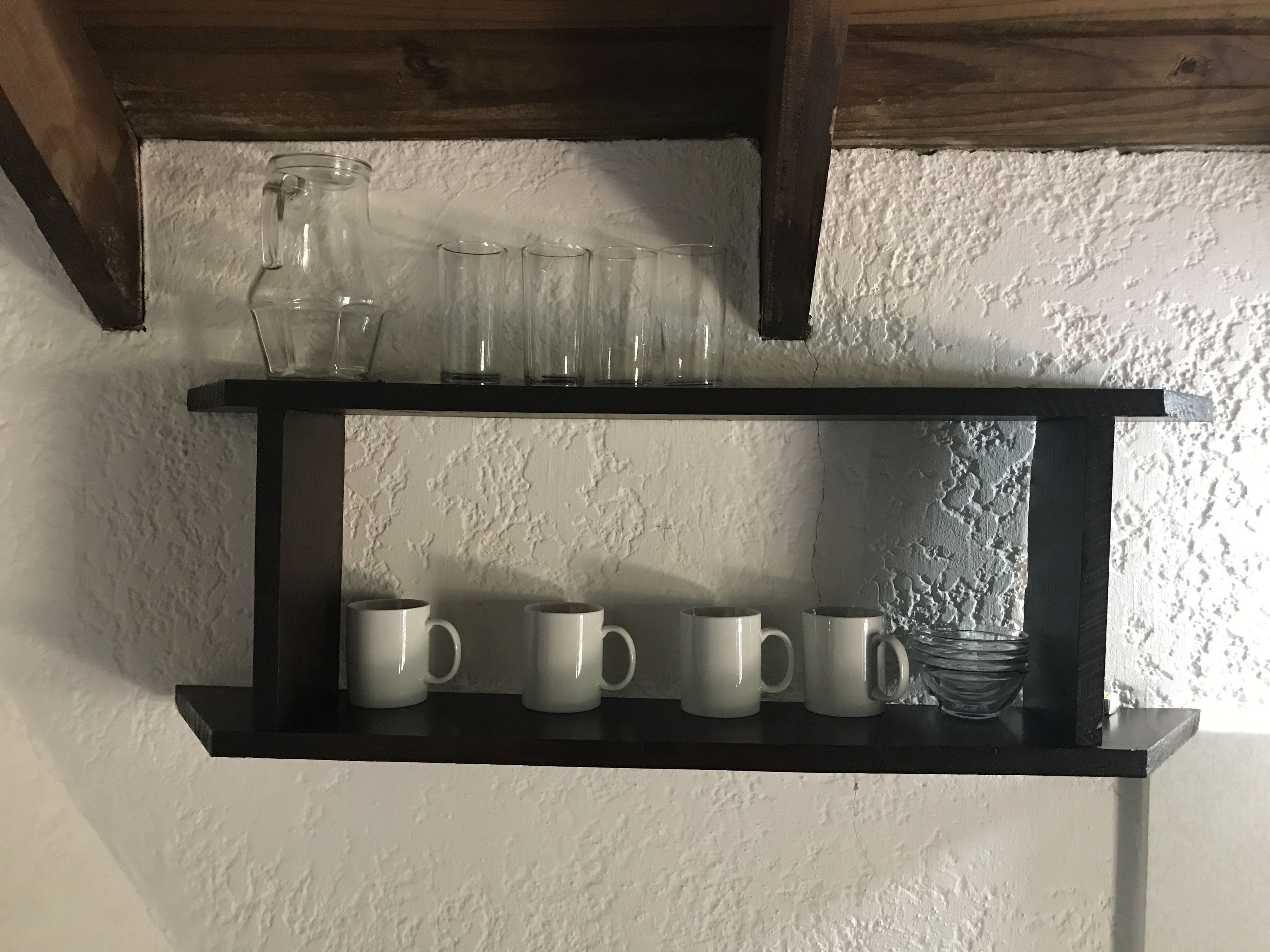 A black shelf with white mugs and a glass jar against a textured wall.
