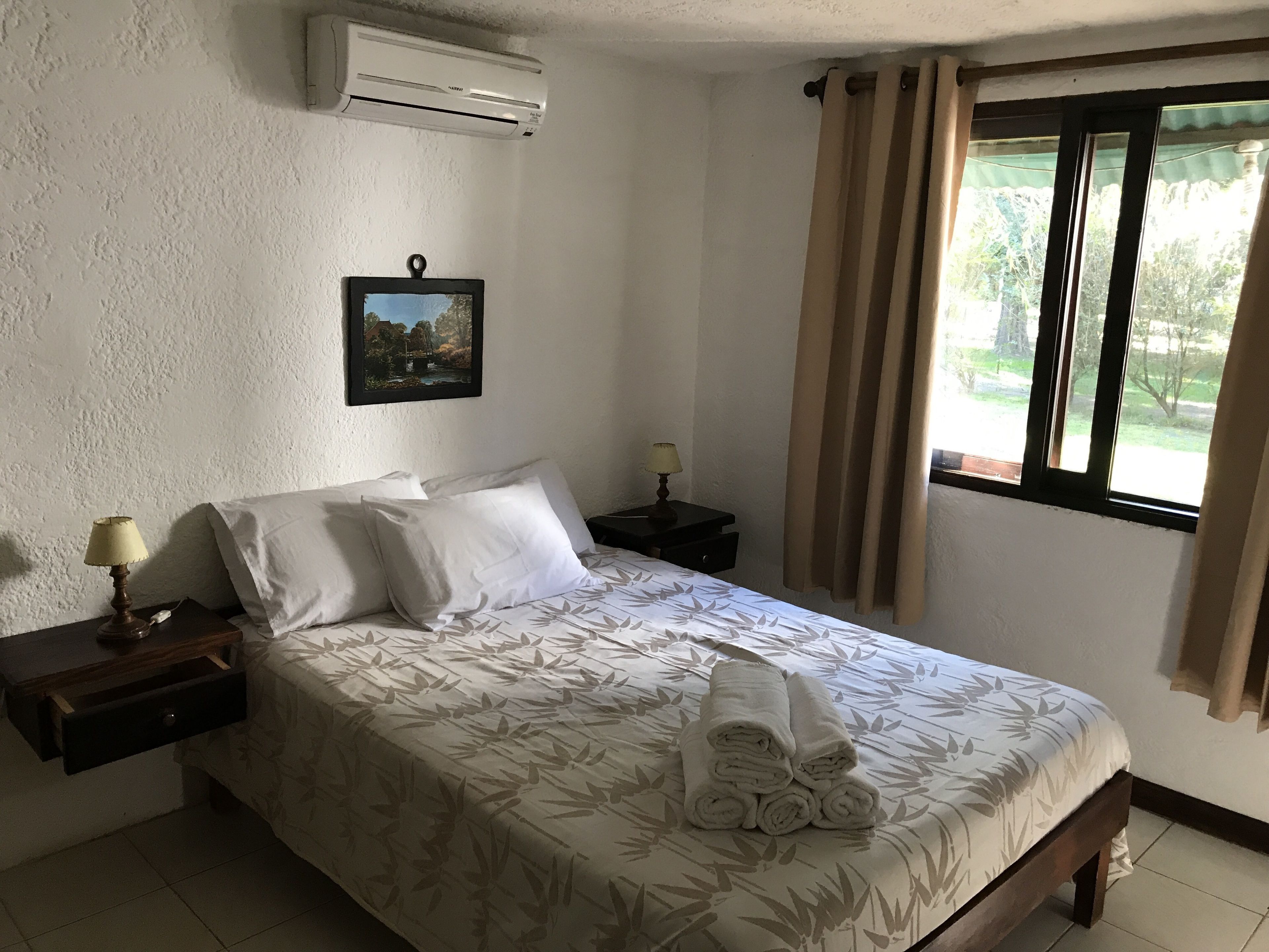 Un lit bien fait, recouvert d’une couvre-lit à motifs floraux, un tableau accroché au mur et un appareil de climatisation placé au-dessus.