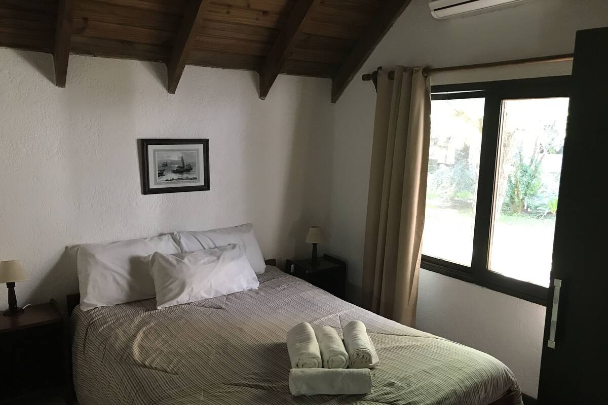 A bedroom with a bed, a nightstand, a lamp, a framed picture on the wall, and a window with curtains.