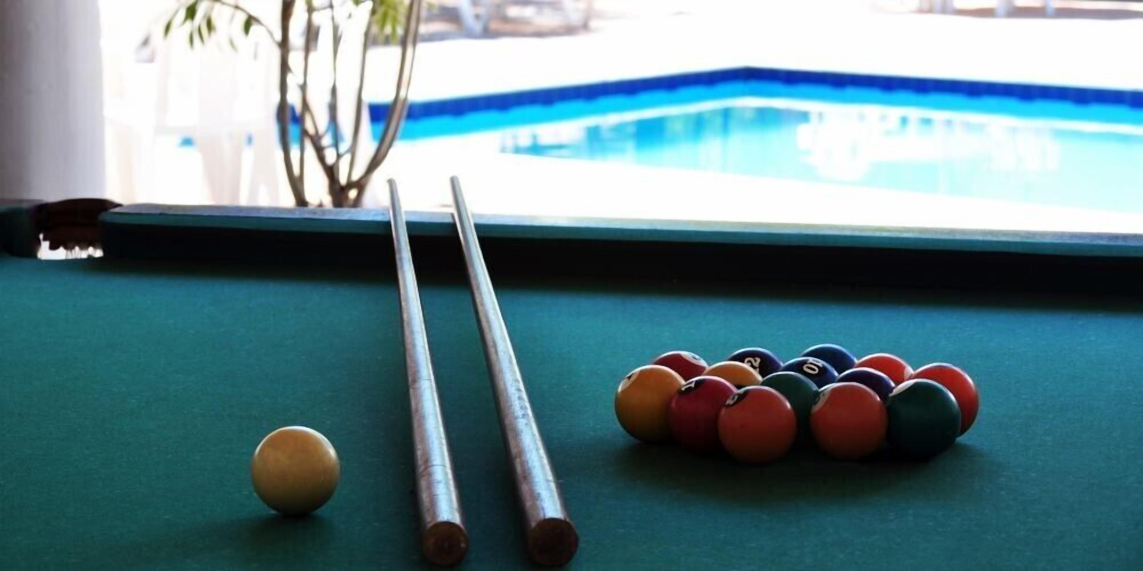 A pool table with billiard balls and cues, a window with a view of a pool, and a plant in the background.