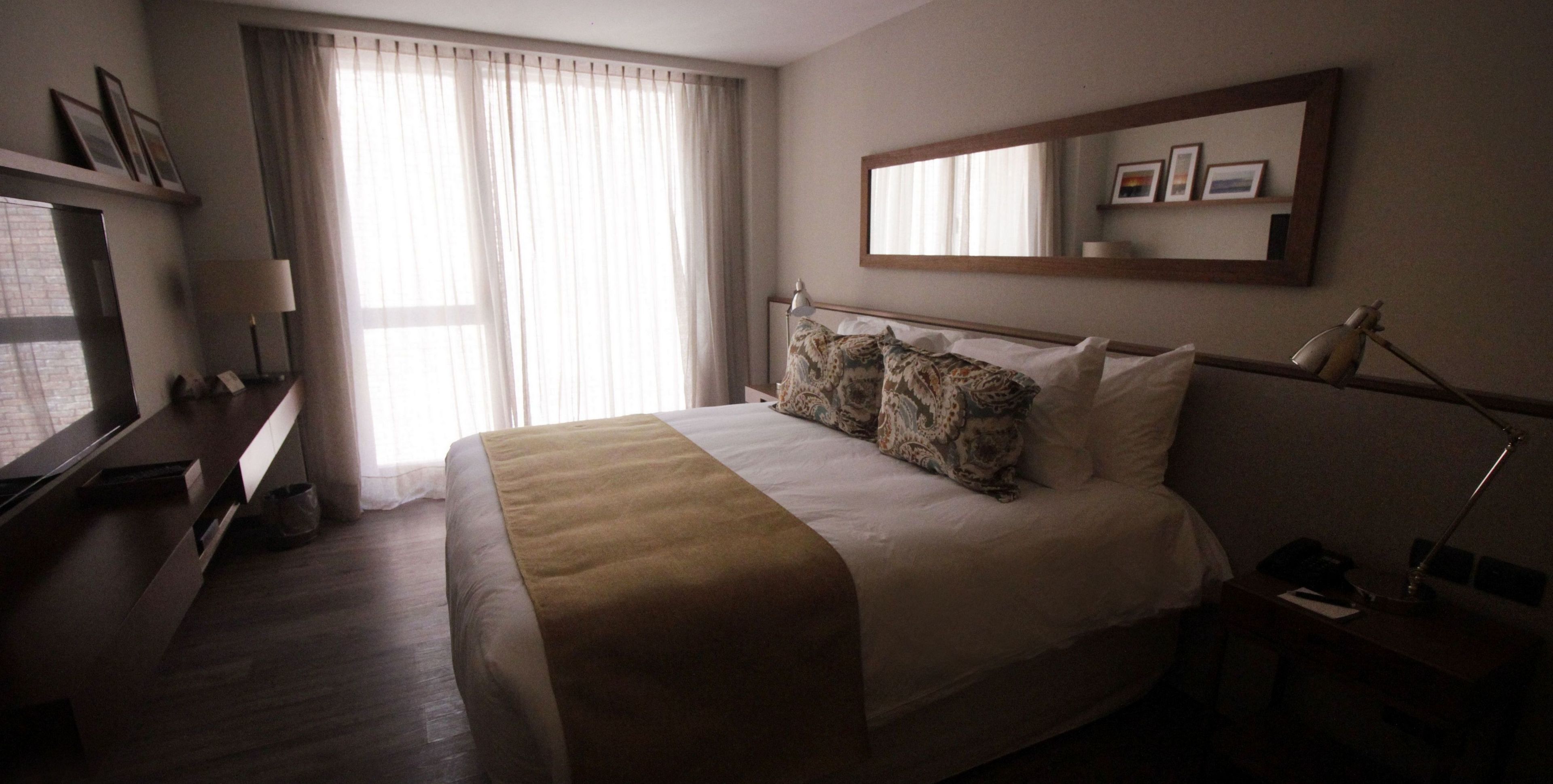 A hotel room with a bed, a desk, a chair, a lamp, a mirror, and a window with curtains.