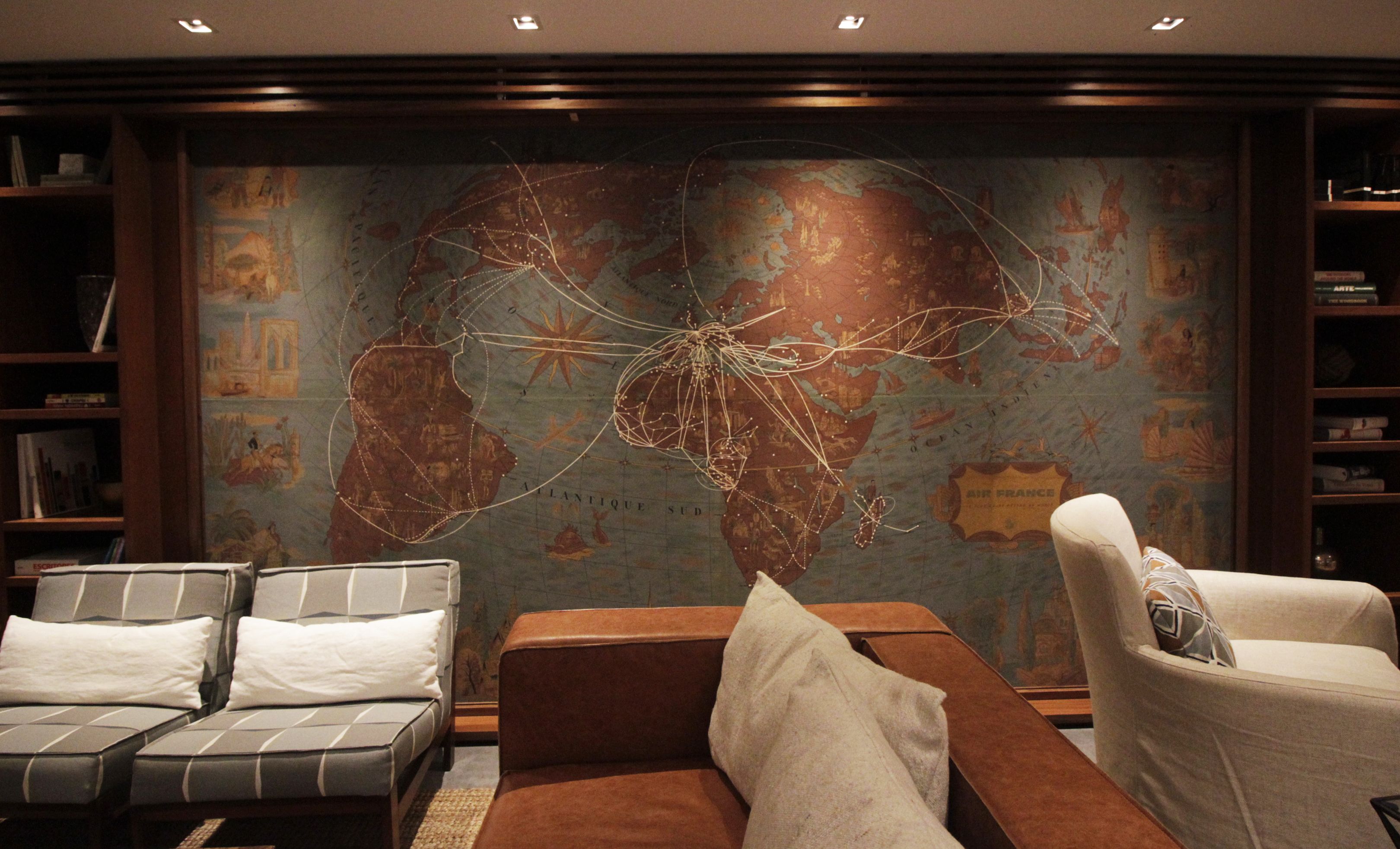 A room with a large world map mural, a brown sofa, a white armchair, and a bookshelf.