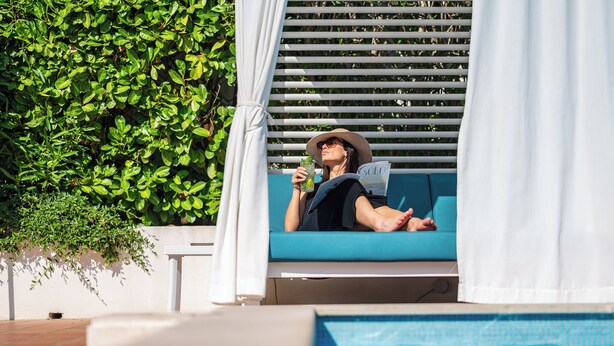 Una persona relajándose en una silla azul junto a una piscina, sosteniendo una revista.