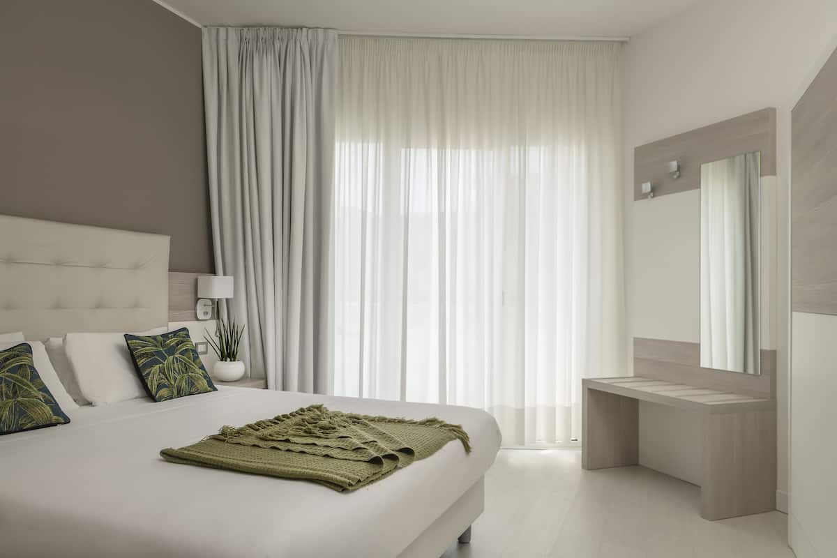 Un dormitorio moderno con una cama grande, una mesita de noche, un espejo y una ventana con cortinas translúcidas.