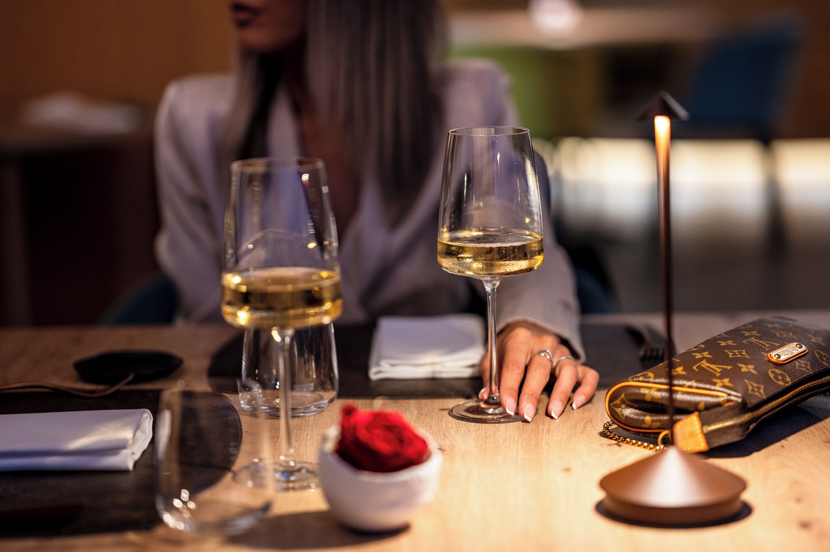 A table with two wine glasses, a candle, a small bowl with a red rose, and a handbag.