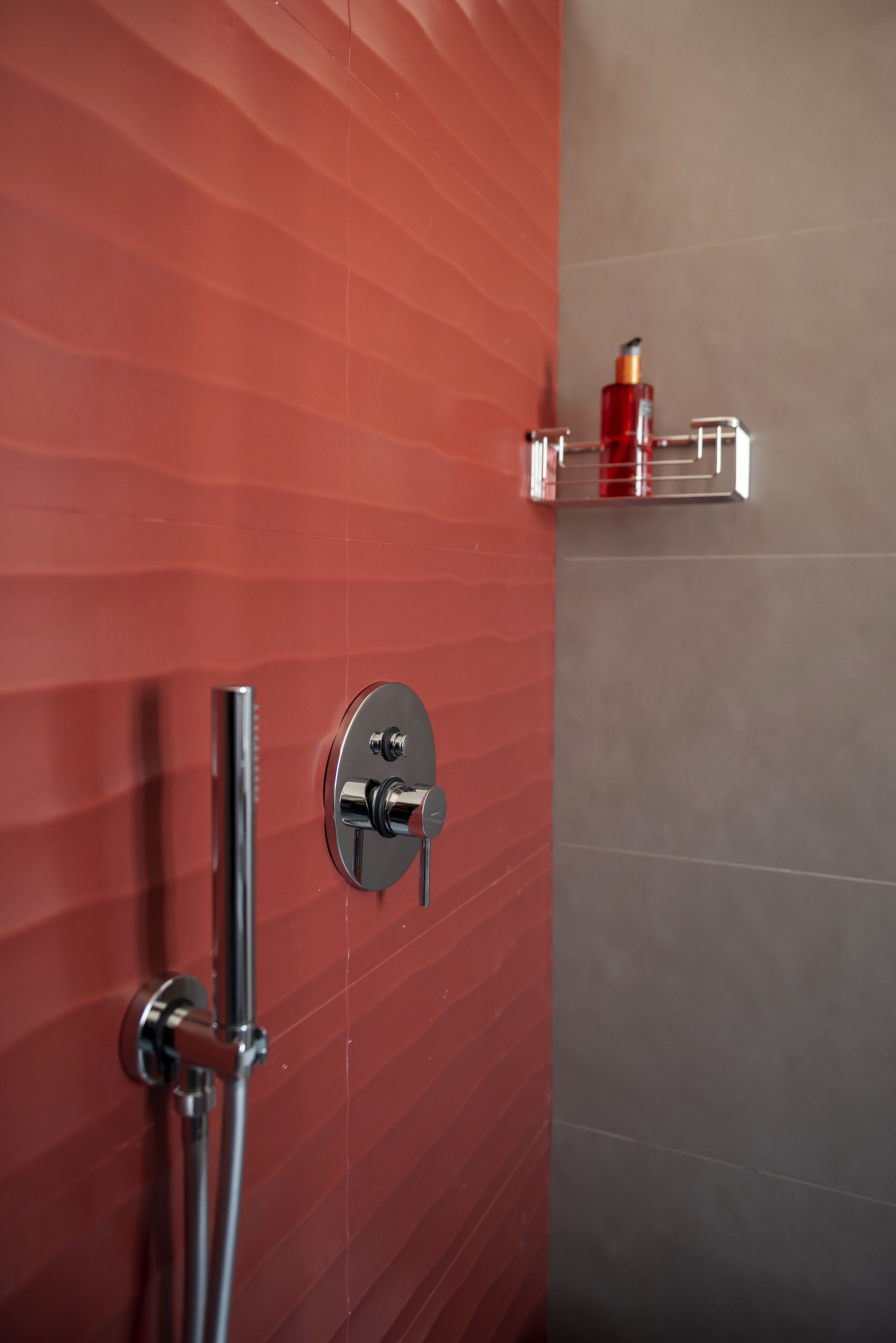 A bathroom with a red tiled wall, a chrome showerhead, and a small shelf with a bottle.