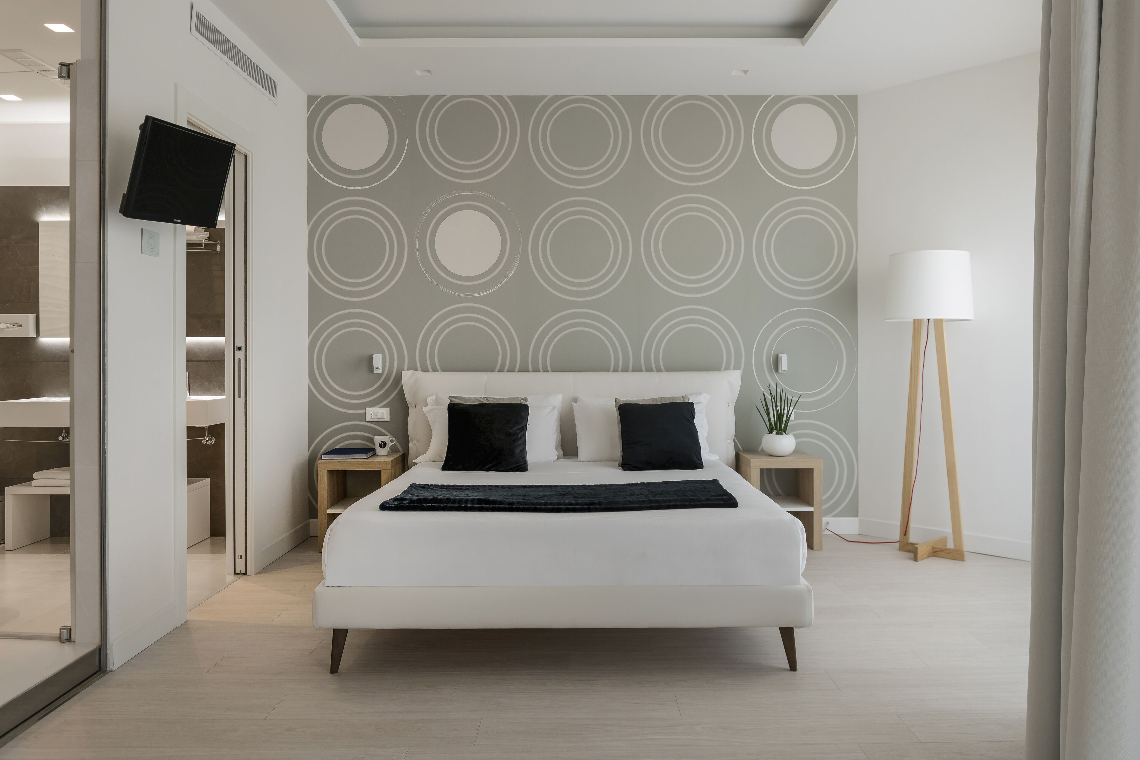 A modern bedroom with a large bed, a flat-screen TV mounted on the wall, and a bedside table with a lamp.