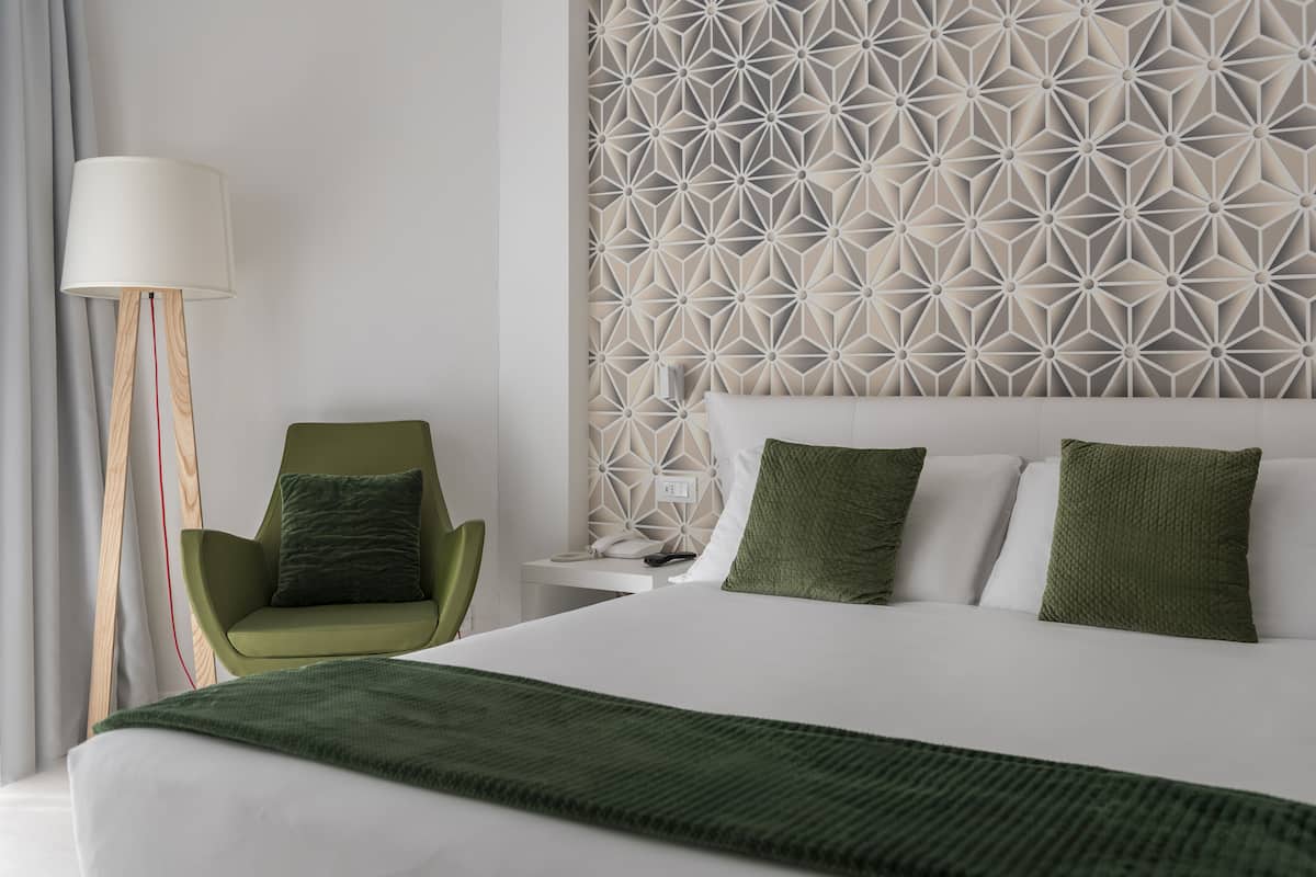 Una habitación de hotel moderna con una cama grande, un sillón verde, una mesita de noche con una lámpara y una pared con un estampado geométrico.