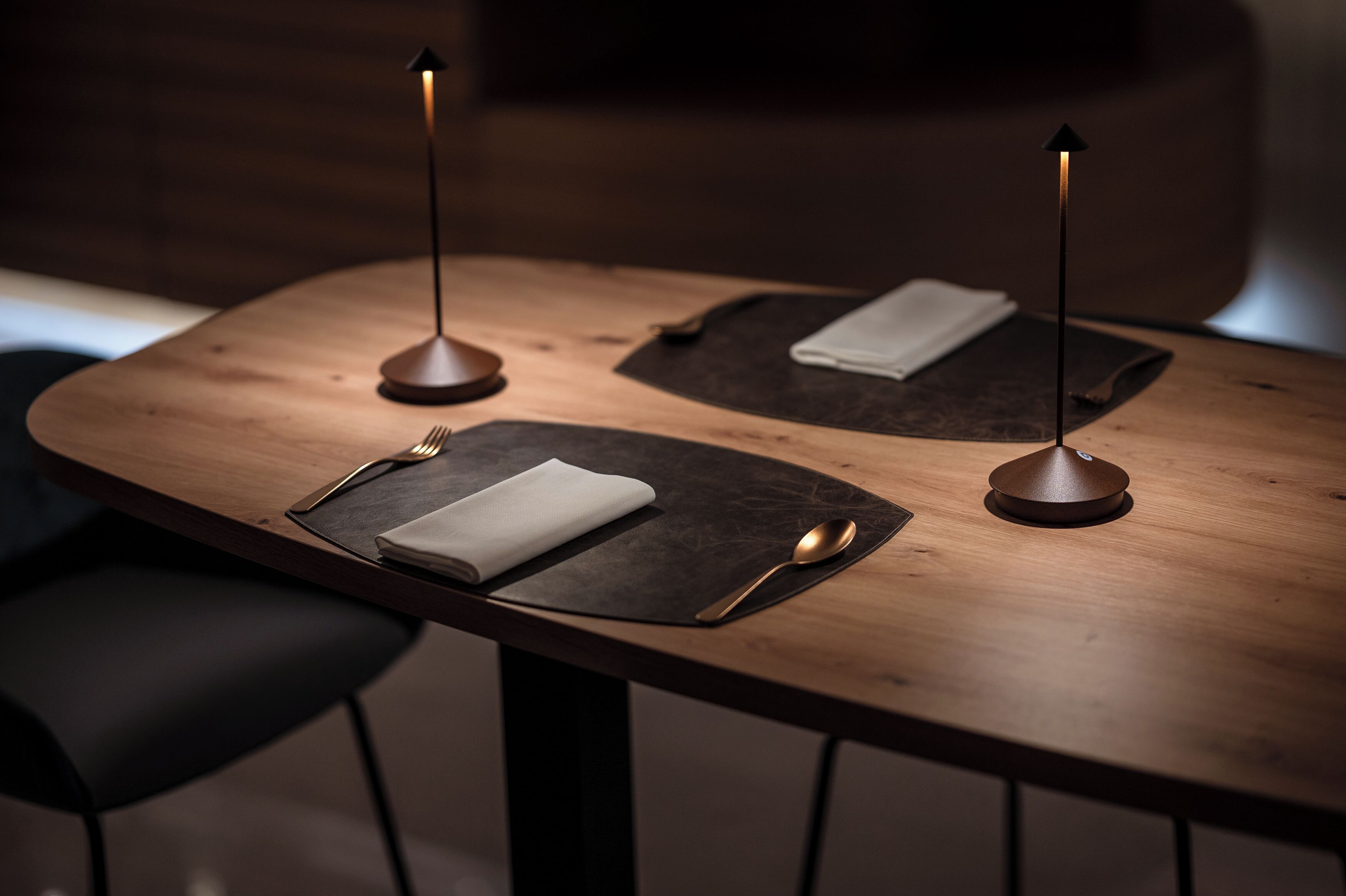 A wooden dining table with three black placemats, each with a folded white napkin and a spoon.