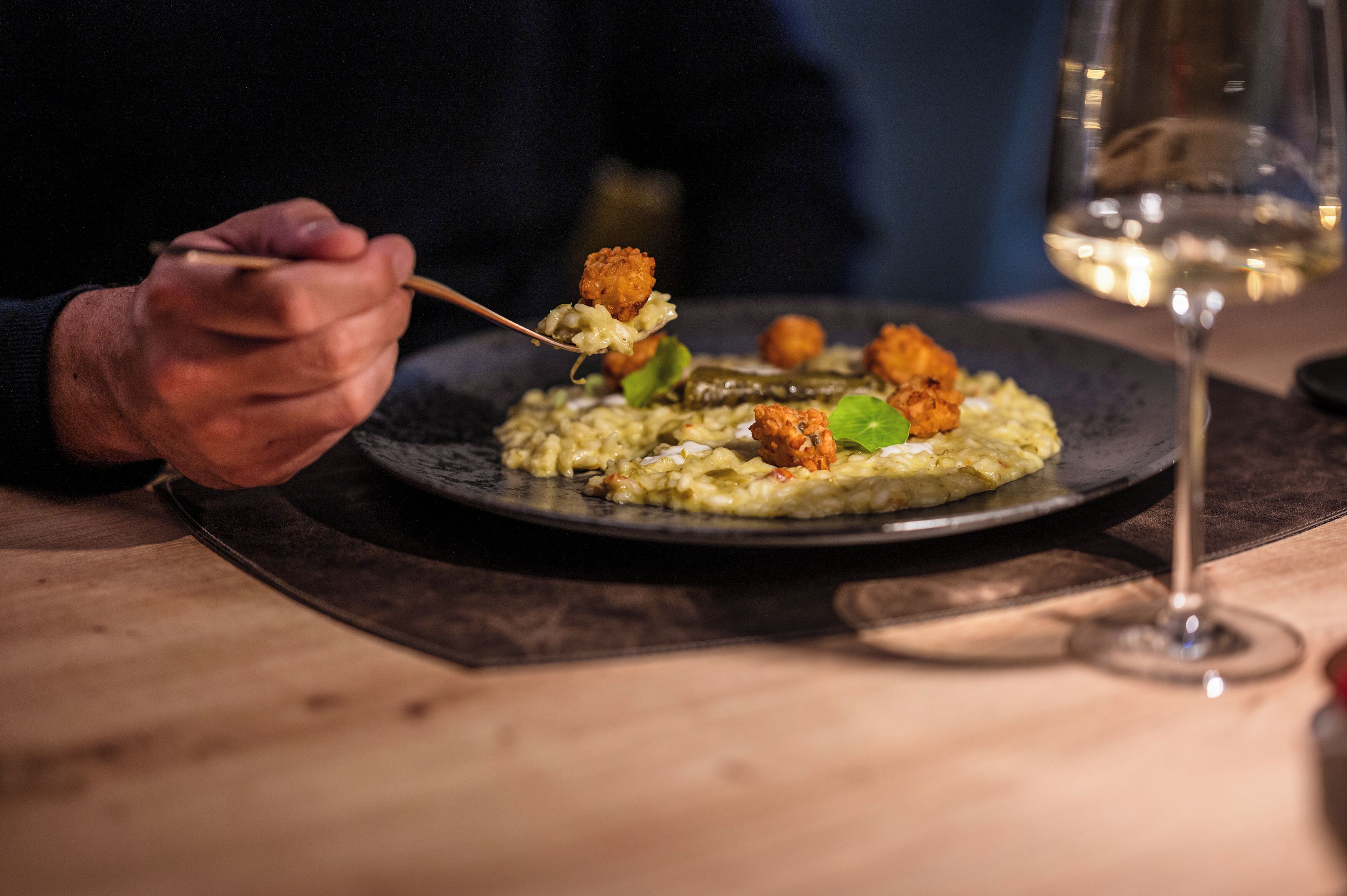 Une personne tenant une fourchette avec de la nourriture dessus, un verre de vin blanc et une assiette de nourriture sur une table.