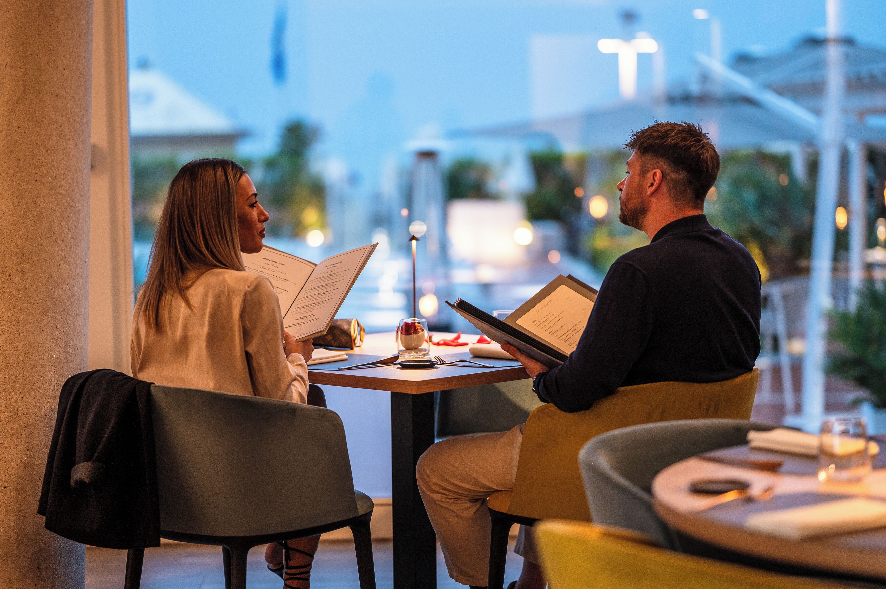 Two people seated at a table with menus, in a well-lit indoor setting with a view of the cityscape outside.