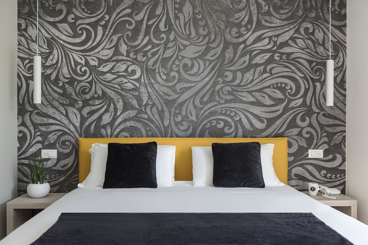 Una cama bien hecha con edredón blanco y negro, cabecera amarilla y dos almohadas negras. Hay una mesita de noche blanca con una planta y una taza de café blanca. La pared tiene un papel tapiz floral gris.