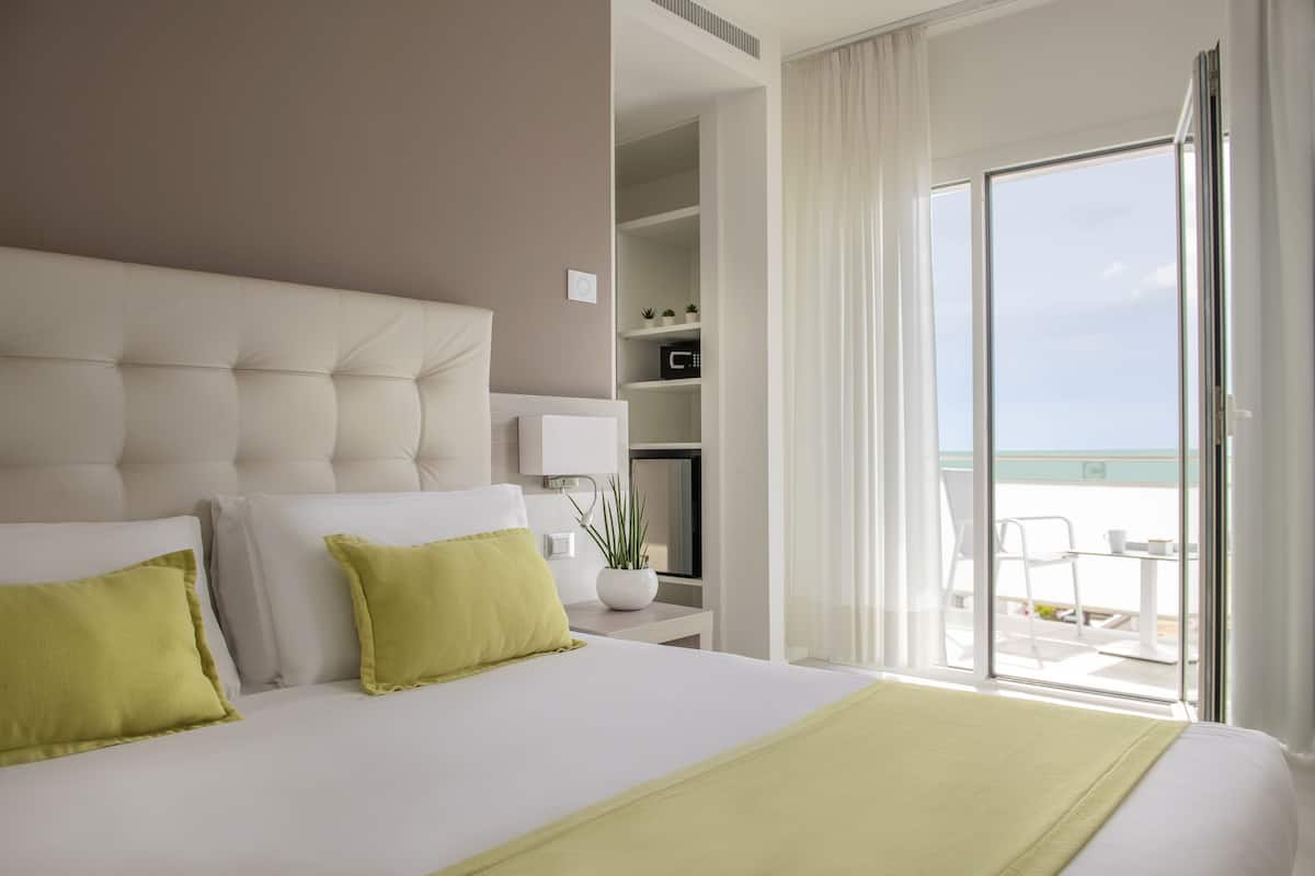 Habitación de hotel con una cama grande, una mesita de noche con lámpara, una planta pequeña en maceta y vista al océano desde las puertas del balcón.