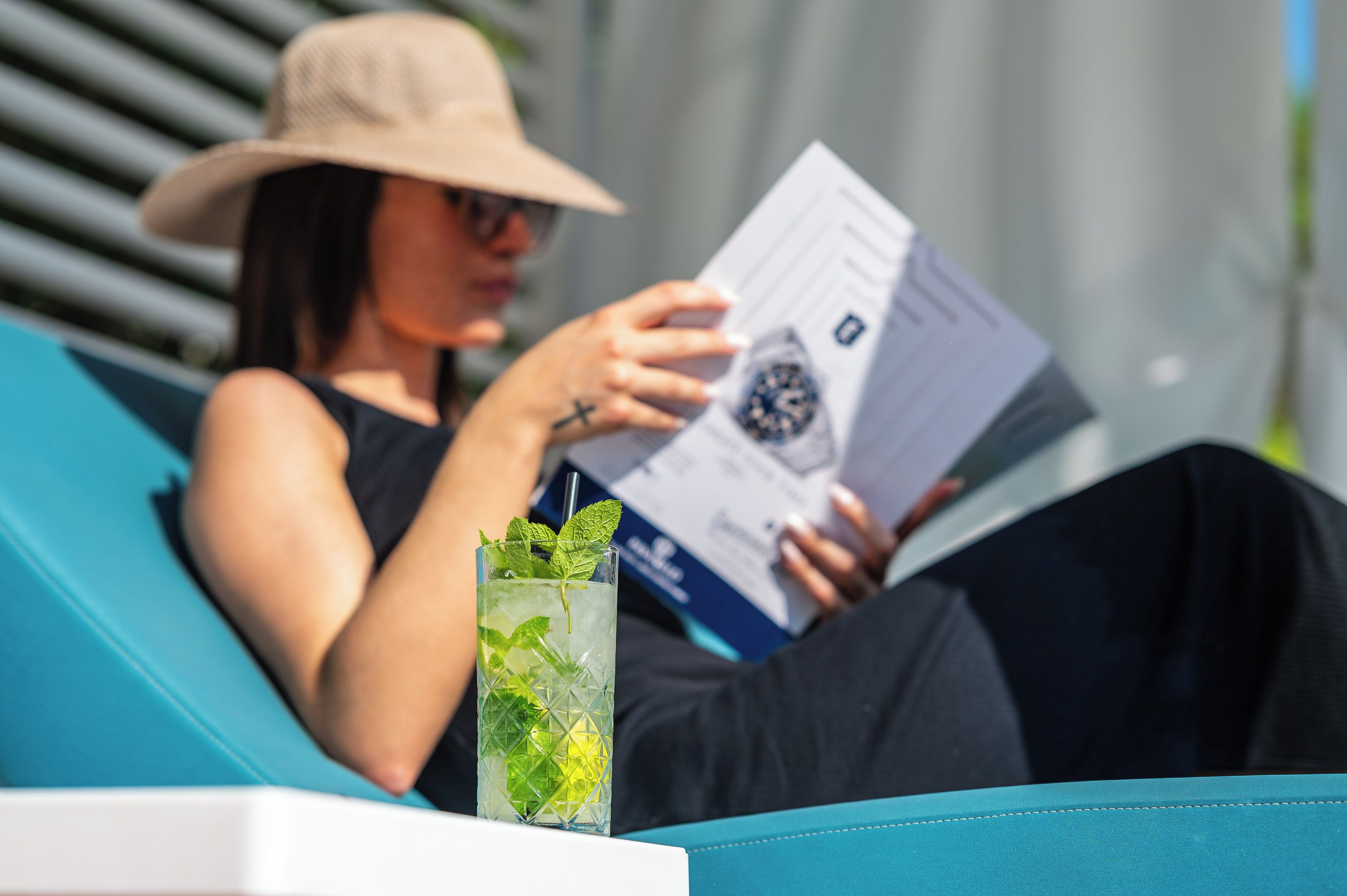 A person sitting on a blue lounge chair reading a magazine, with a glass of iced drink with mint on the table.